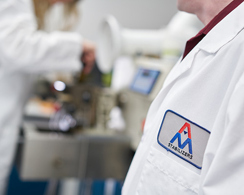 Close up of person wearing a lab coat with AM Stabilizers badge, blurred person with lab coat working in the bckground